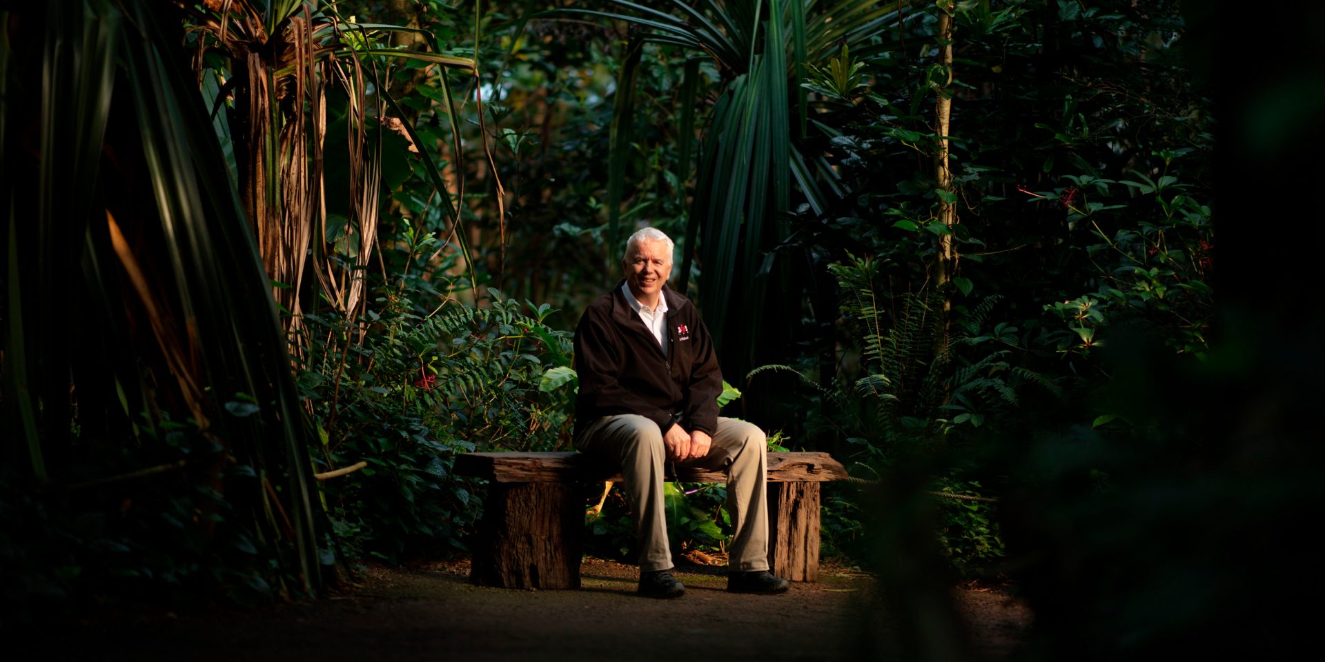 Zoodirektor Alex Rübel sitzt auf einer Holzbank in der Masoalahalle. Im Hintergrund sind die Bäume des Dschungels sichtbar.