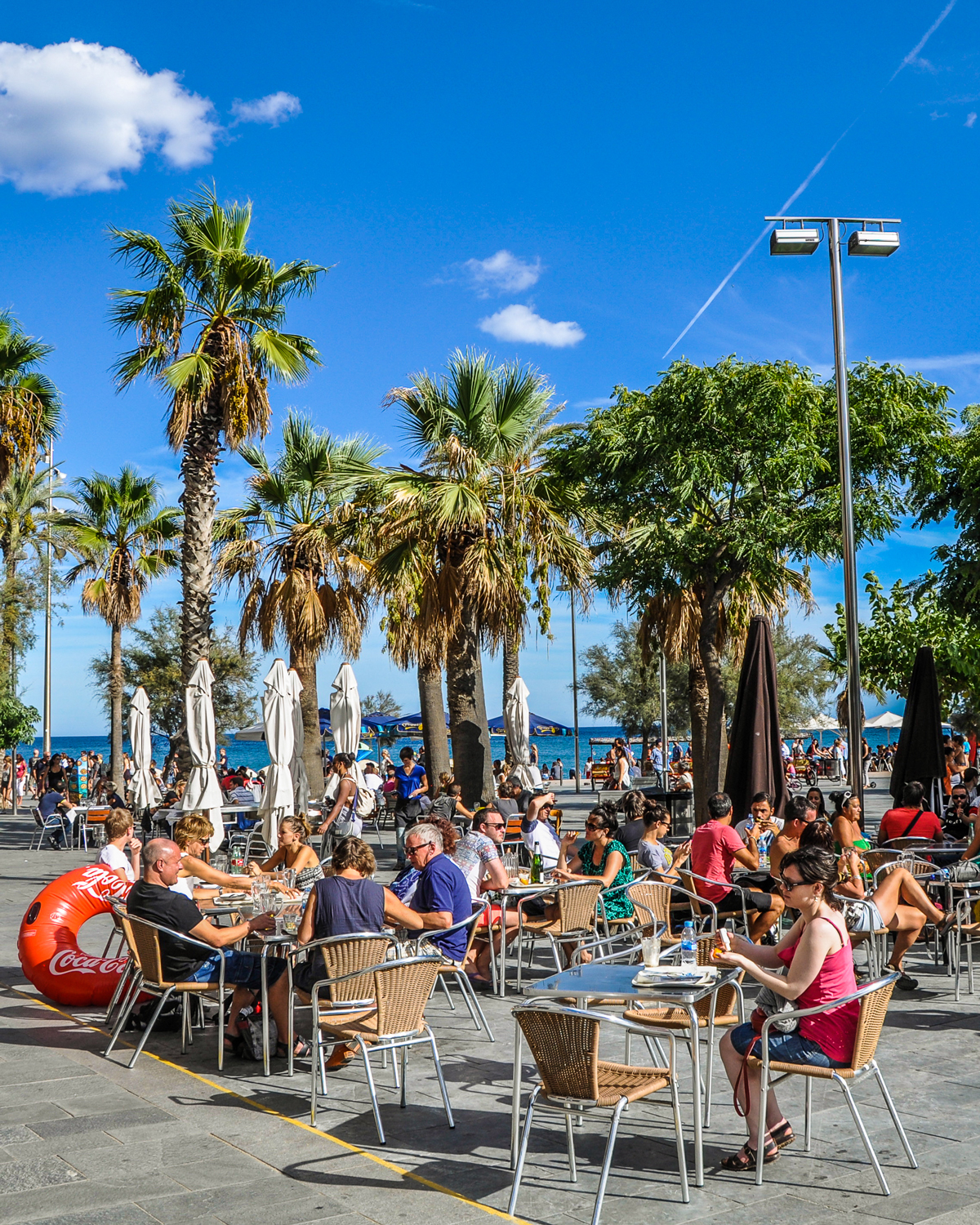 Menschen sitzen draussen an Tischen an der Strandpromenade mit Palmen im Hintergrund