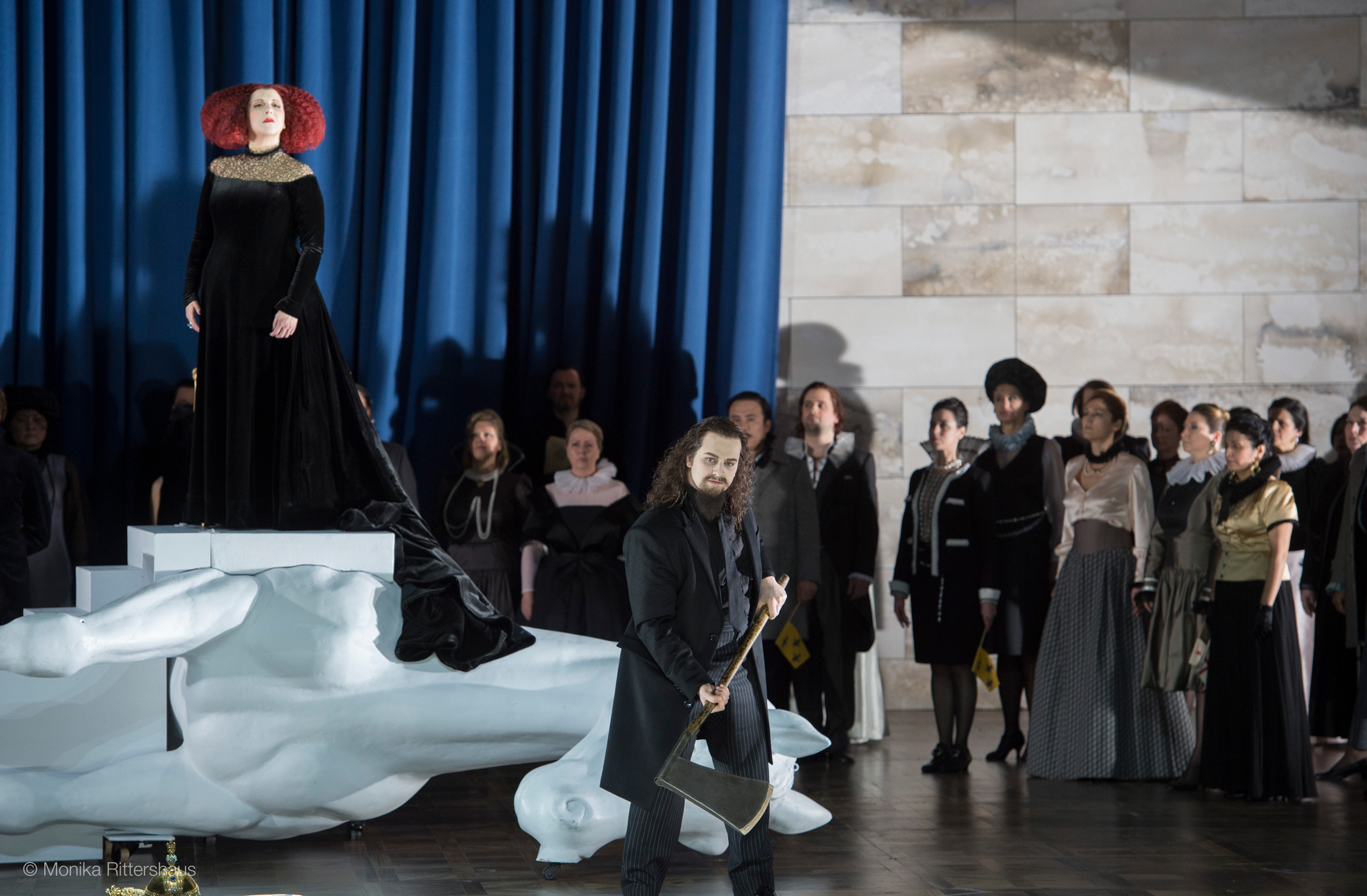 Solist der Aufführung Maria Stuarda mit einer Axt in der Hand. Im Hintergrund stehen die Solistin und der Chor.
