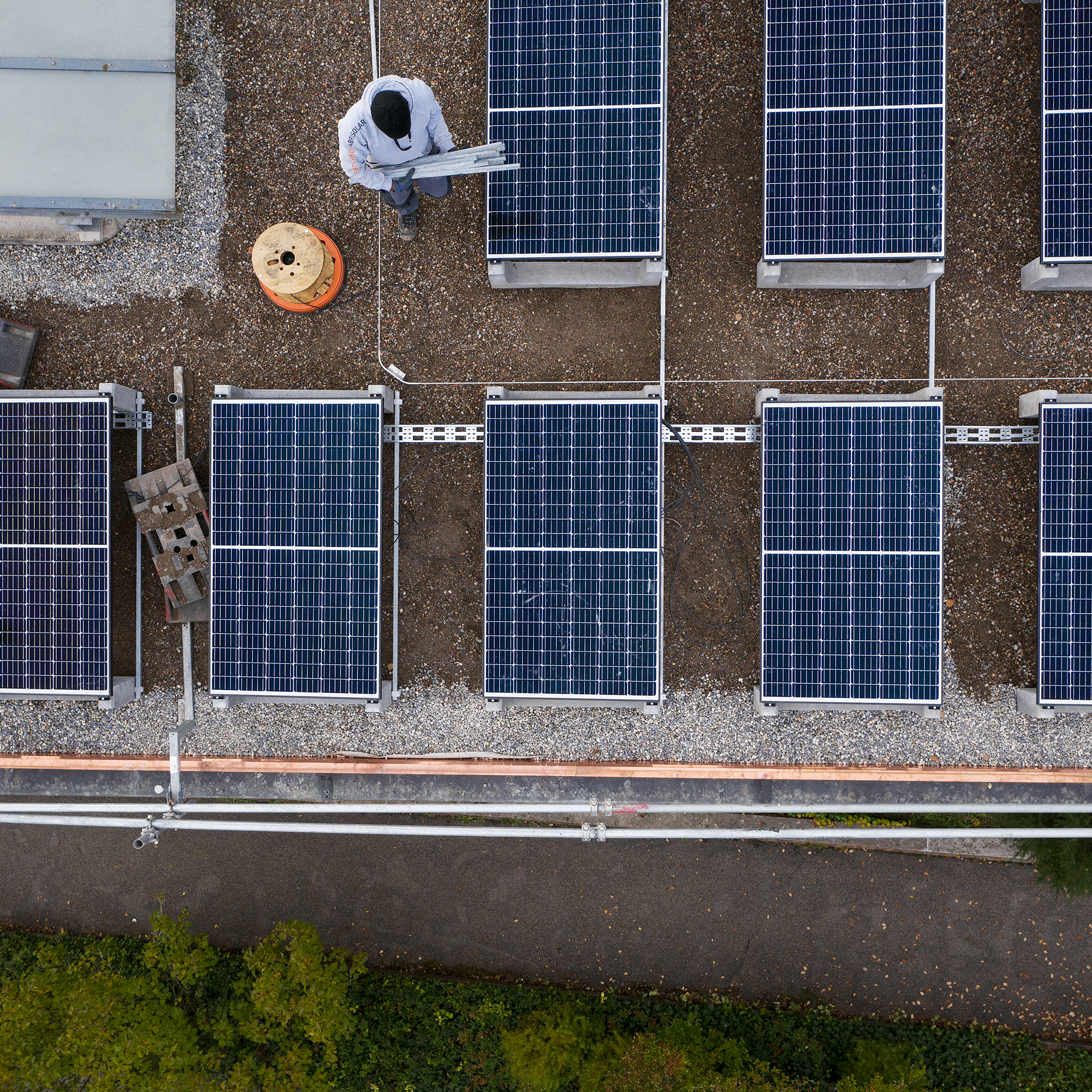 Drohenaufnahme, die neun Solarpannels auf dem Schulhausdach zeigt. Dazwischen bewegt sich ein Arbeiter. Auch ihn sieht man von oben.