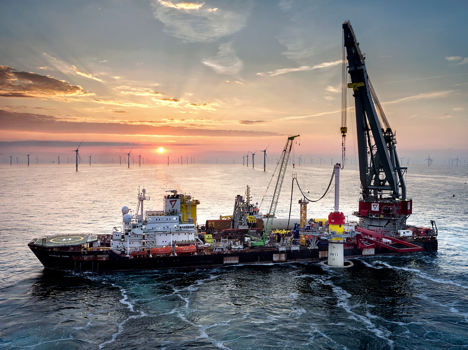 Ein Schiff, das für den Bau der Windturbinen verwendet wird, steht im Vordergrund. Im Hintergrund viele Windturbinen im Meer. Es ist Sonnenuntergang.