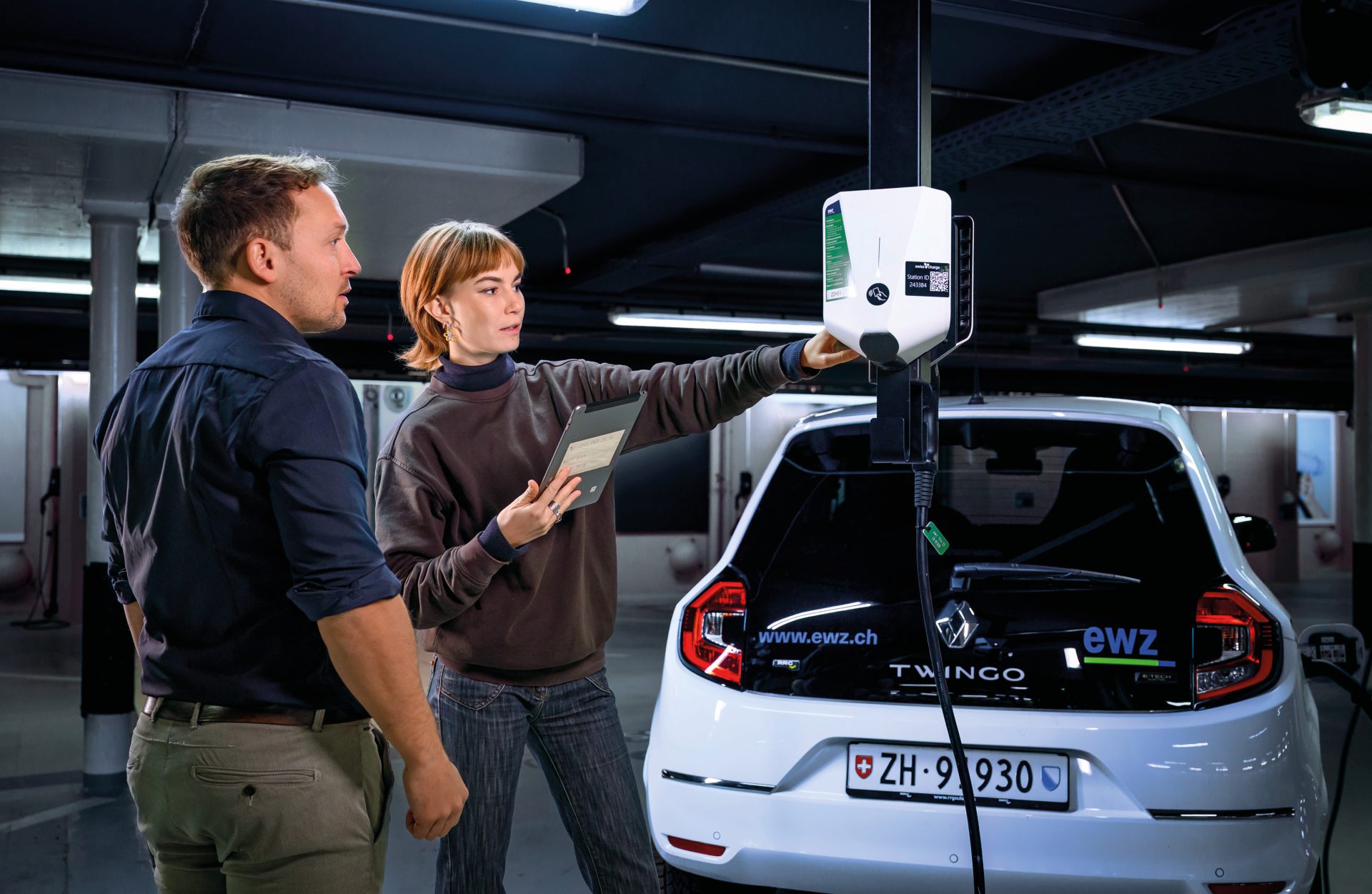 Zwei Fachpersonen konfigurieren eine smarte Ladestation für ein Elektrofahrzeug in einer Tiefgarage – die Lösung trägt zur netzverträglichen Steuerung von Stromflüssen bei und unterstützt die stabile Einbindung von Elektromobilität ins Stromnetz für eine zuverlässige Energieversorgung.
